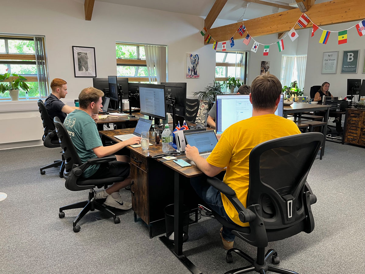 Open-plan office with several employees working at desks equipped with computers and laptops in a bright workspace with exposed wooden beams, large windows, indoor plants, and international flags hanging from the ceiling.