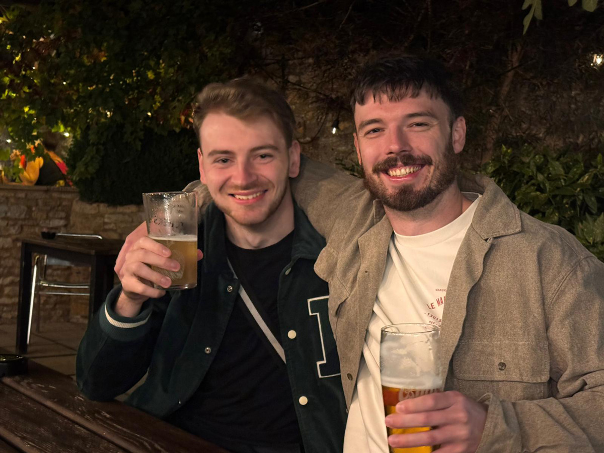 . Two men seated at a table, each holding a beer, engaged in conversation. 