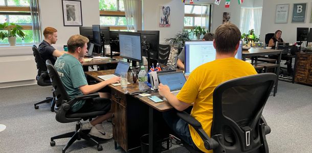 Open-plan office with several employees working at desks equipped with computers and laptops in a bright workspace with exposed wooden beams, large windows, indoor plants, and international flags hanging from the ceiling.