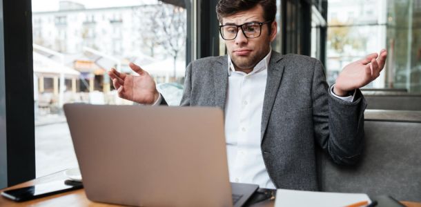Confused businessman in eyeglasses sitting by the table in cafe while shrugs his shoulders and looking at laptop computer